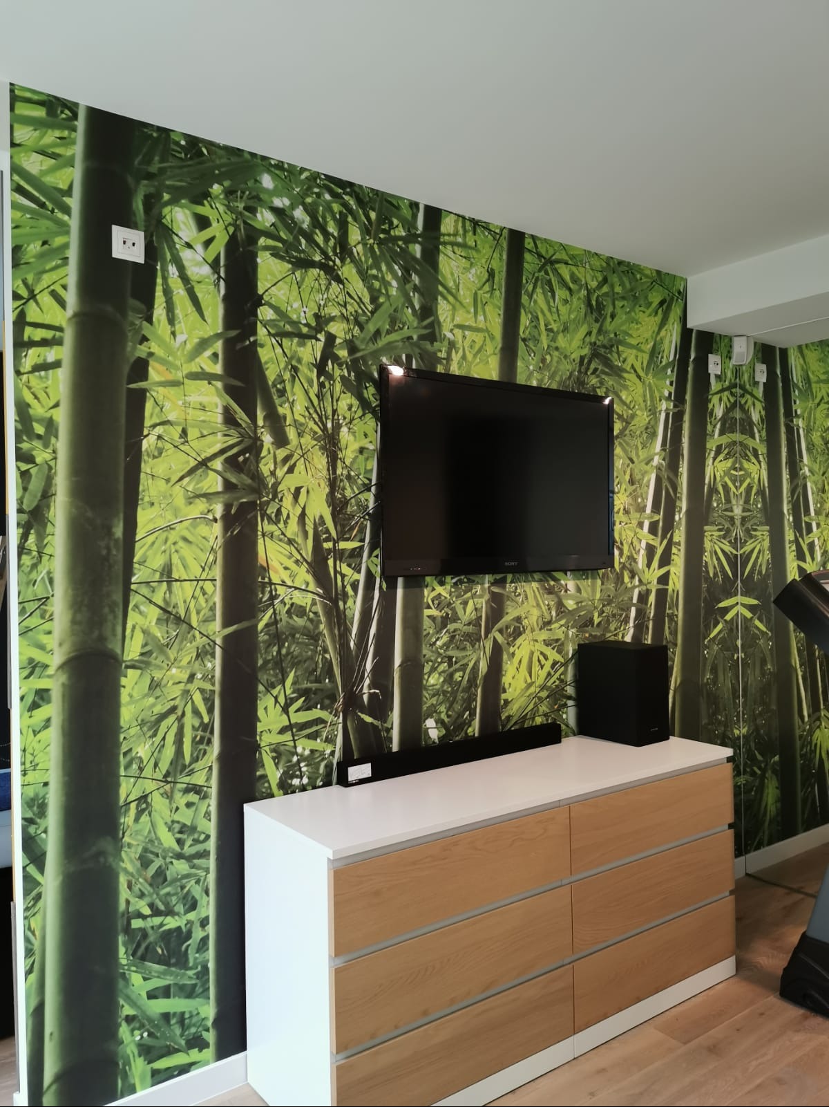 Pokój z fototapetą bambusową, szafka RTV z białymi bokami i drewnianymi frontami, telewizor na ścianie. Nowoczesne wnętrze z akcentem natury.