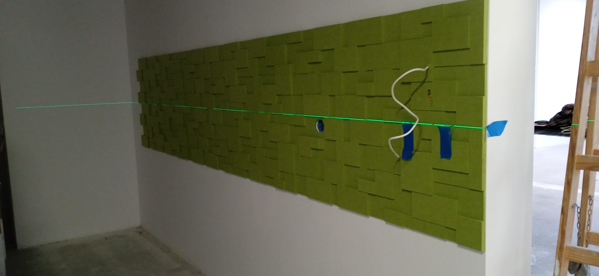 Montaż dekoracyjnych paneli ściennych 3D w kolorze zielonym, z użyciem poziomicy laserowej. Widoczne kable i taśmy montażowe, co sugeruje etap instalacji.