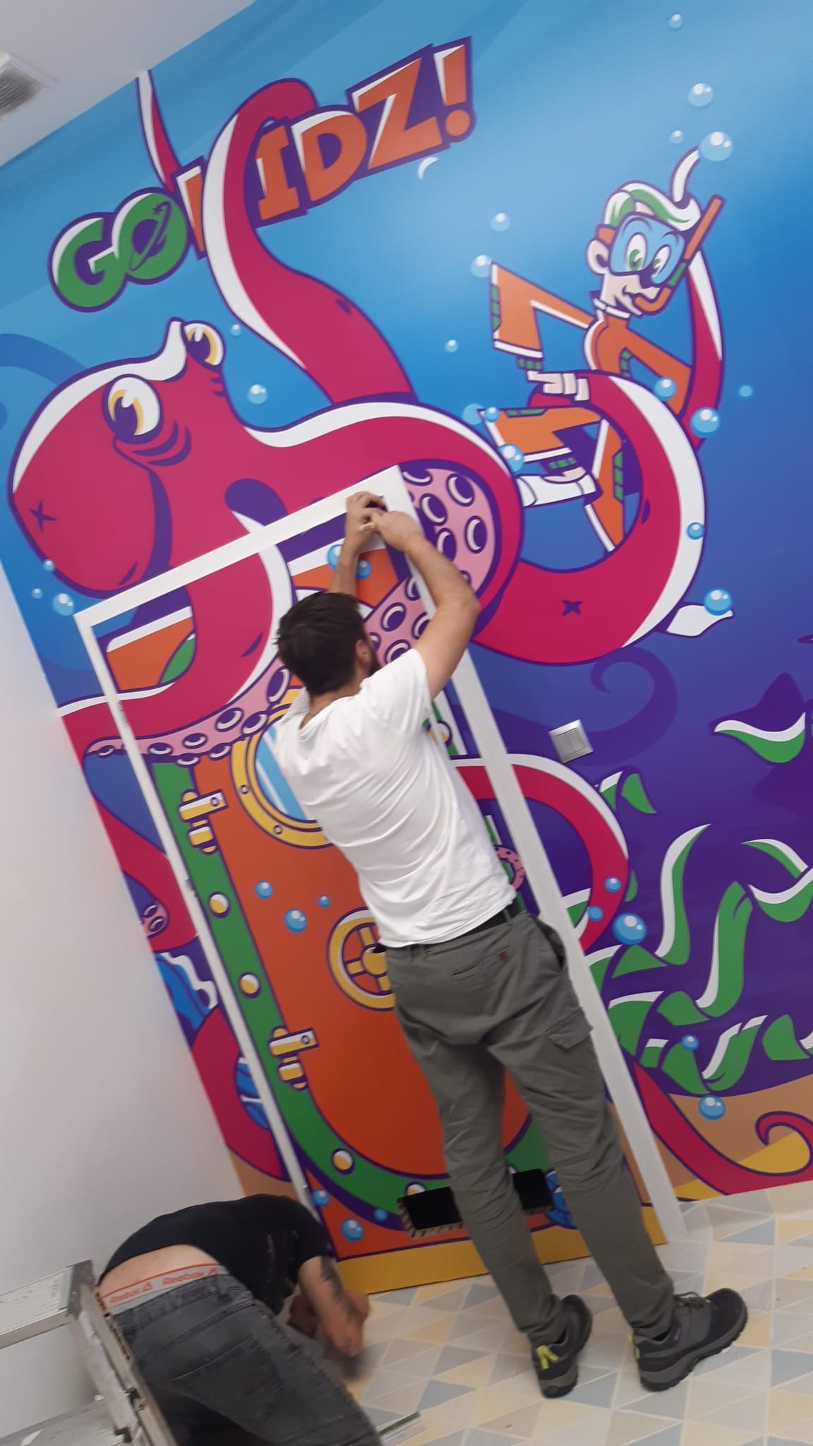 Montaż kolorowej grafiki z motywem morskim na ścianie w pokoju dziecięcym. Dwóch mężczyzn przy pracy, jeden na drabinie, drugi na podłodze. Widoczne detale grafiki: ośmiornica, nurek.