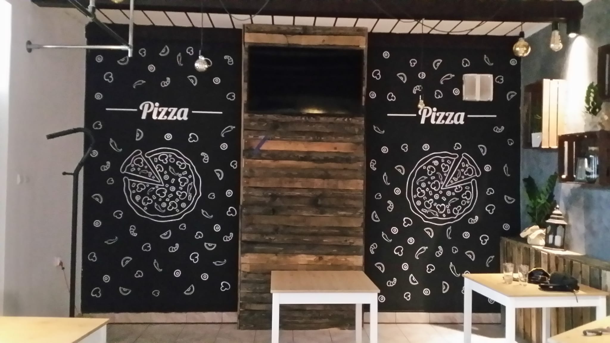 Ściana w pizzerii z rysunkami pizzy kredą na czarnej farbie tablicowej, drewniane panele i stoliki. Dekoracja wnętrza lokalu gastronomicznego.