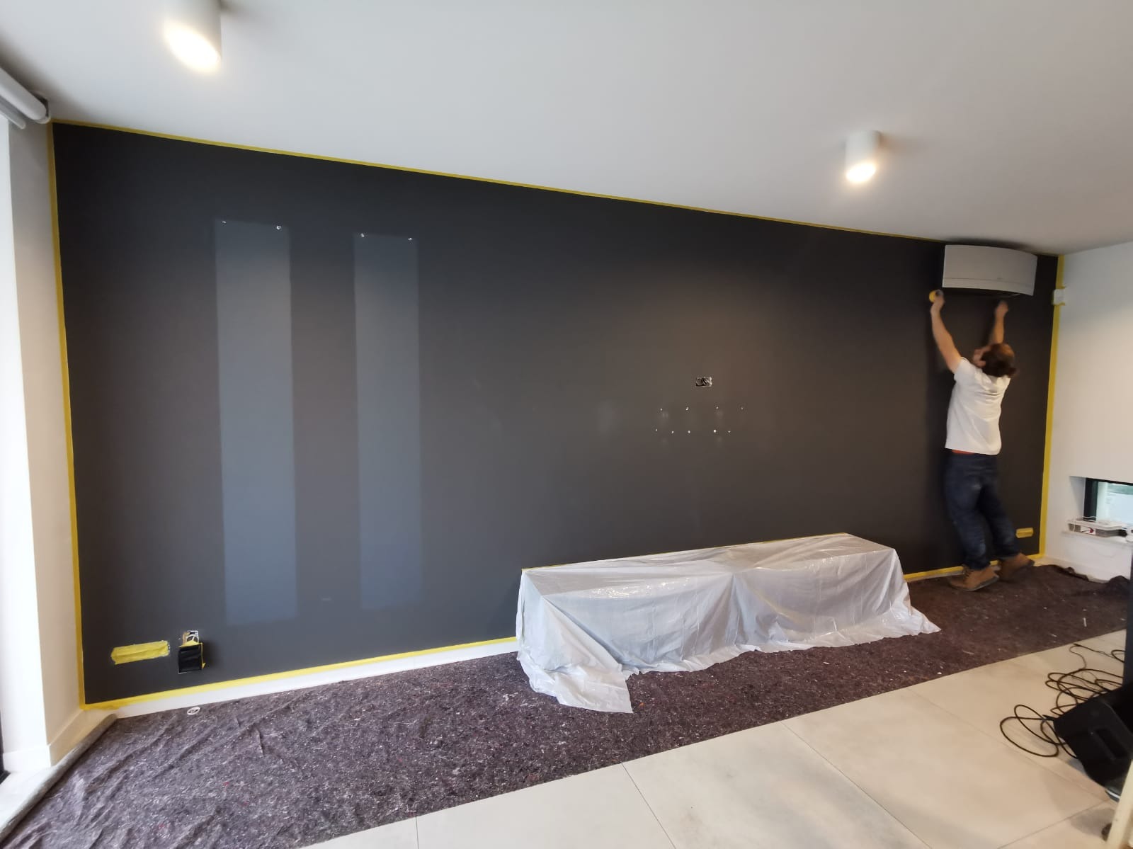Malowanie ściany na ciemny kolor w pokoju. Mężczyzna maluje górną część ściany przy klimatyzatorze. Podłoga i meble zabezpieczone folią i taśmą malarską.