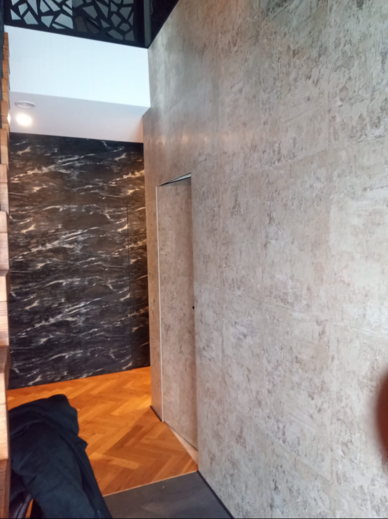 Wykończone wnętrze z panelami ściennymi imitującymi kamień, parkietem w jodełkę i dekoracyjnym sufitem. Widoczny fragment garderoby lub przejścia do innego pomieszczenia.