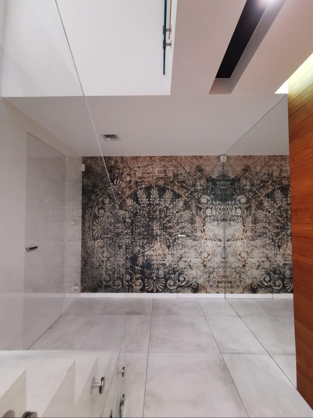 Wnętrze z dekoracyjną tapetą w stylu vintage na ścianie, jasnymi płytkami podłogowymi i minimalistycznymi drzwiami. Widoczne elementy szklane i drewniane.