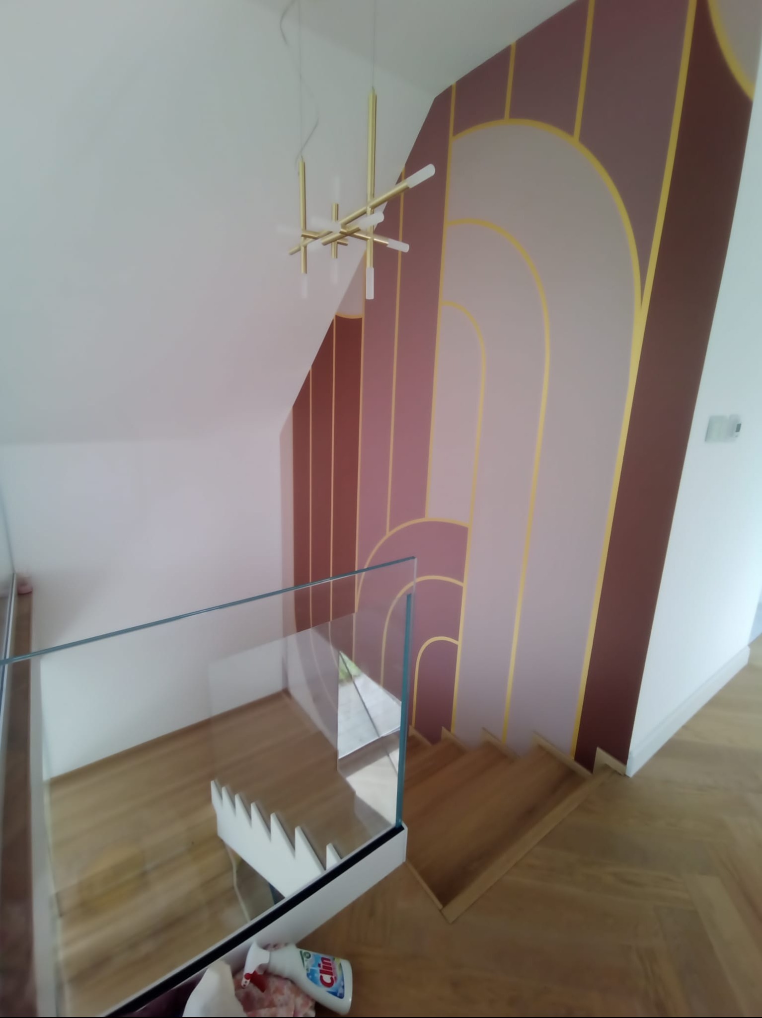 Artystyczne malowanie ściany na klatce schodowej w stylu Art Deco. Geometryczne wzory w odcieniach różu i złota. Nowoczesna lampa wisząca i szklana balustrada.