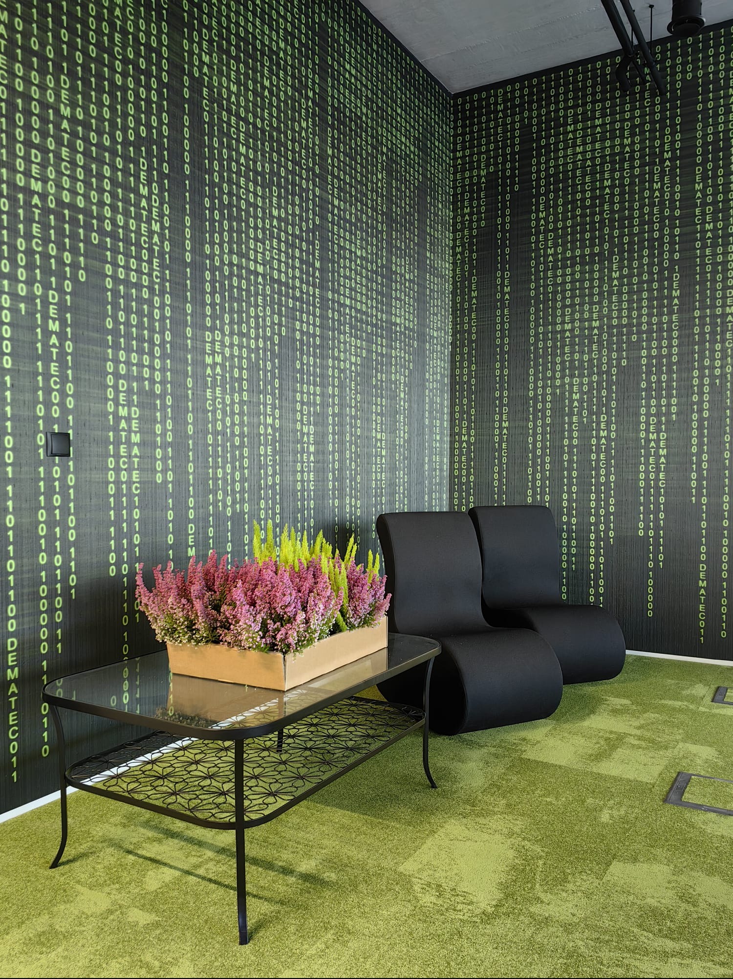 Nowoczesne biuro z tapetą w stylu kodu binarnego, czarne fotele i stolik z kwiatami, tworzące unikalną przestrzeń relaksu.