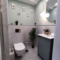 MA DSGN - Kompleksowe remonty i wykończenia - Nowoczesna łazienka w odcieniach szarości i zieleni, z marmurowymi płytkami, wiszącą toaletą, umywalką z szafką w kolorze zielonym, okrągłym lustrem z oświetleniem i dekoracyjnymi obrazkami...