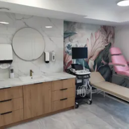 Nowoczesne wnętrze gabinetu lekarskiego z marmurową ścianą, drewnianą szafką z umywalką i sprzętem medycznym, w tym różowym fotelem ginekologicznym.