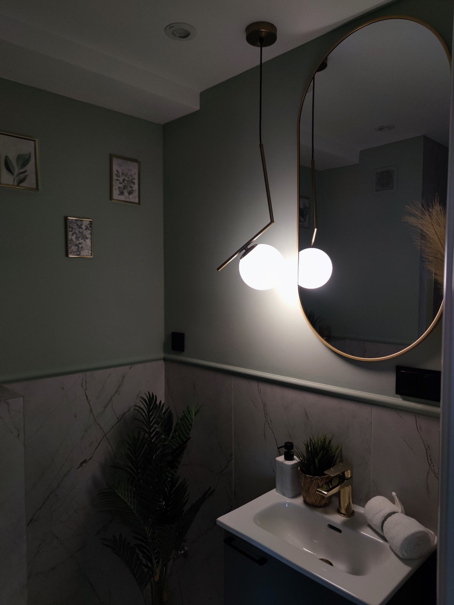 Nowoczesna łazienka w odcieniach zieleni z marmurowymi płytkami, wiszącą lampą z dwoma kulami i złotą armaturą, dekorowana ramkami z grafikami roślinnymi i roślinami doniczkowymi.