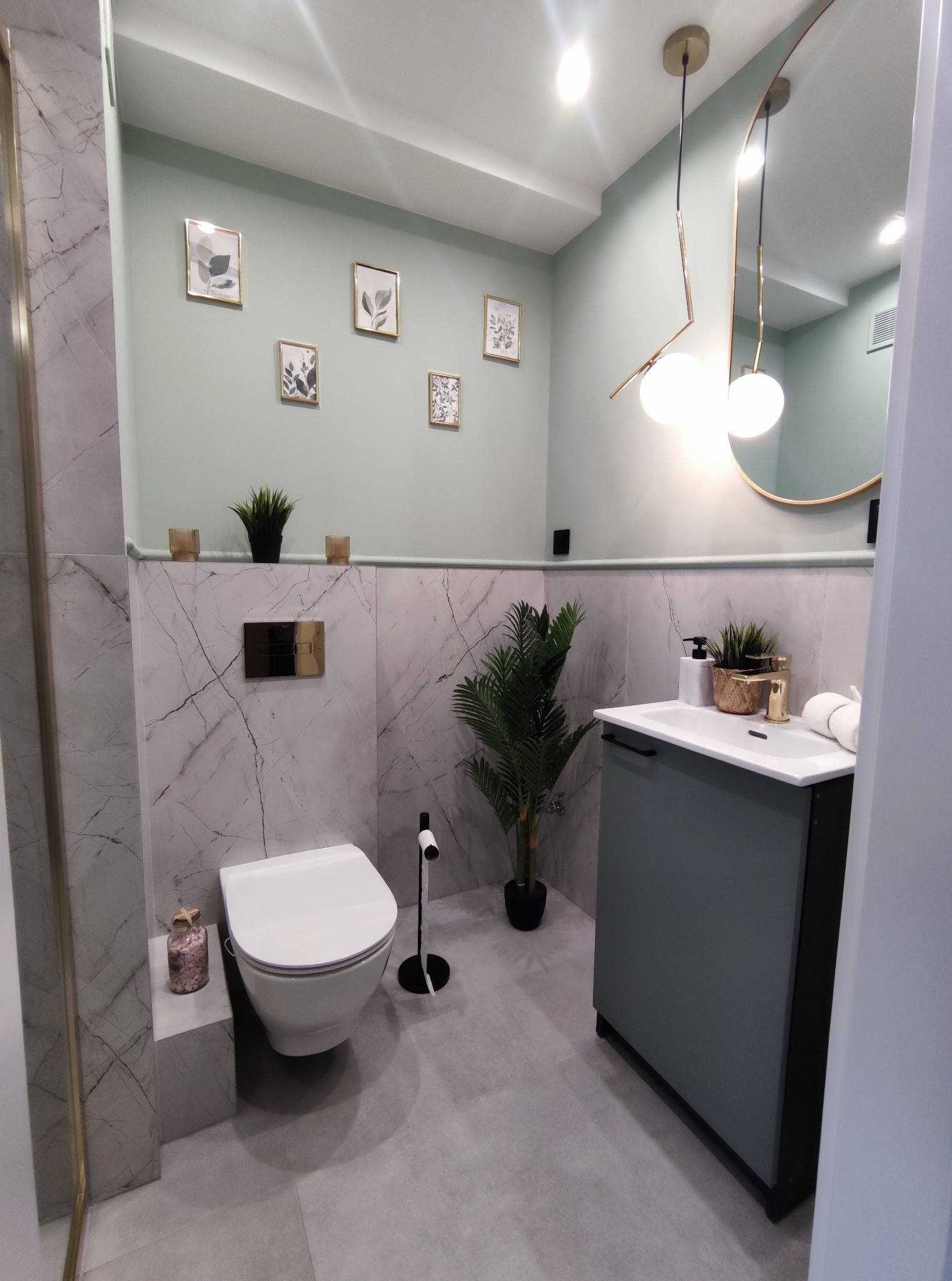 Nowoczesna łazienka w odcieniach szarości i zieleni, z marmurowymi płytkami, wiszącą toaletą, umywalką z szafką w kolorze zielonym, okrągłym lustrem z oświetleniem i dekoracyjnymi obrazkami...