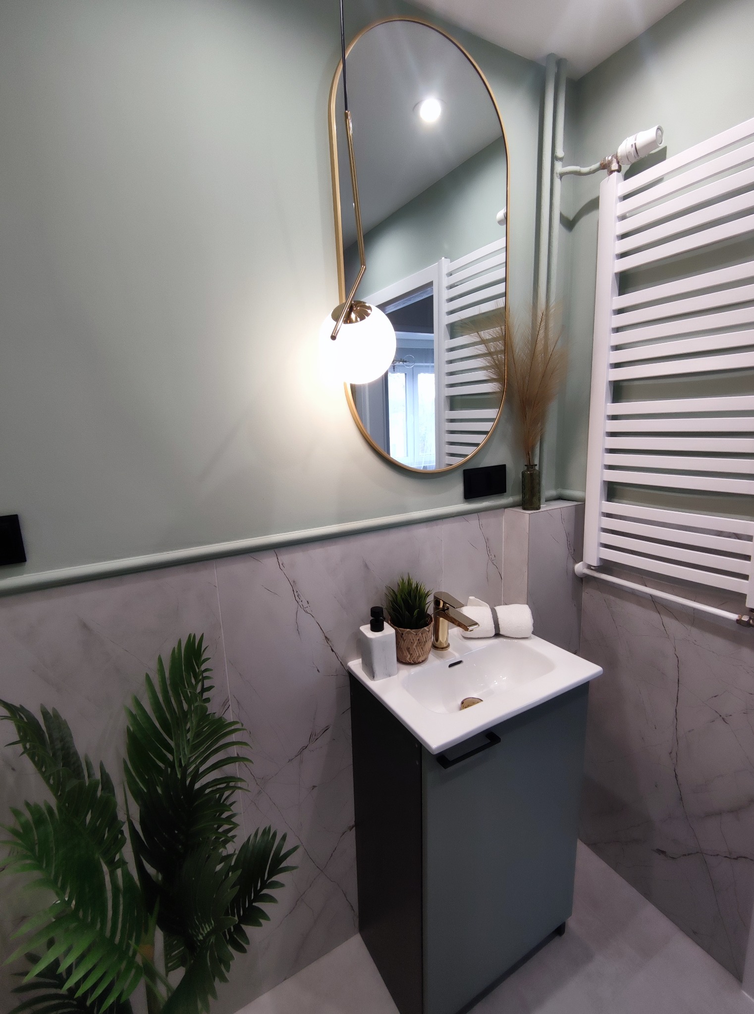 Nowoczesna łazienka w odcieniach szarości i zieleni z marmurowymi płytkami, umywalką z szafką, owalnym lustrem z oświetleniem i grzejnikiem drabinkowym.