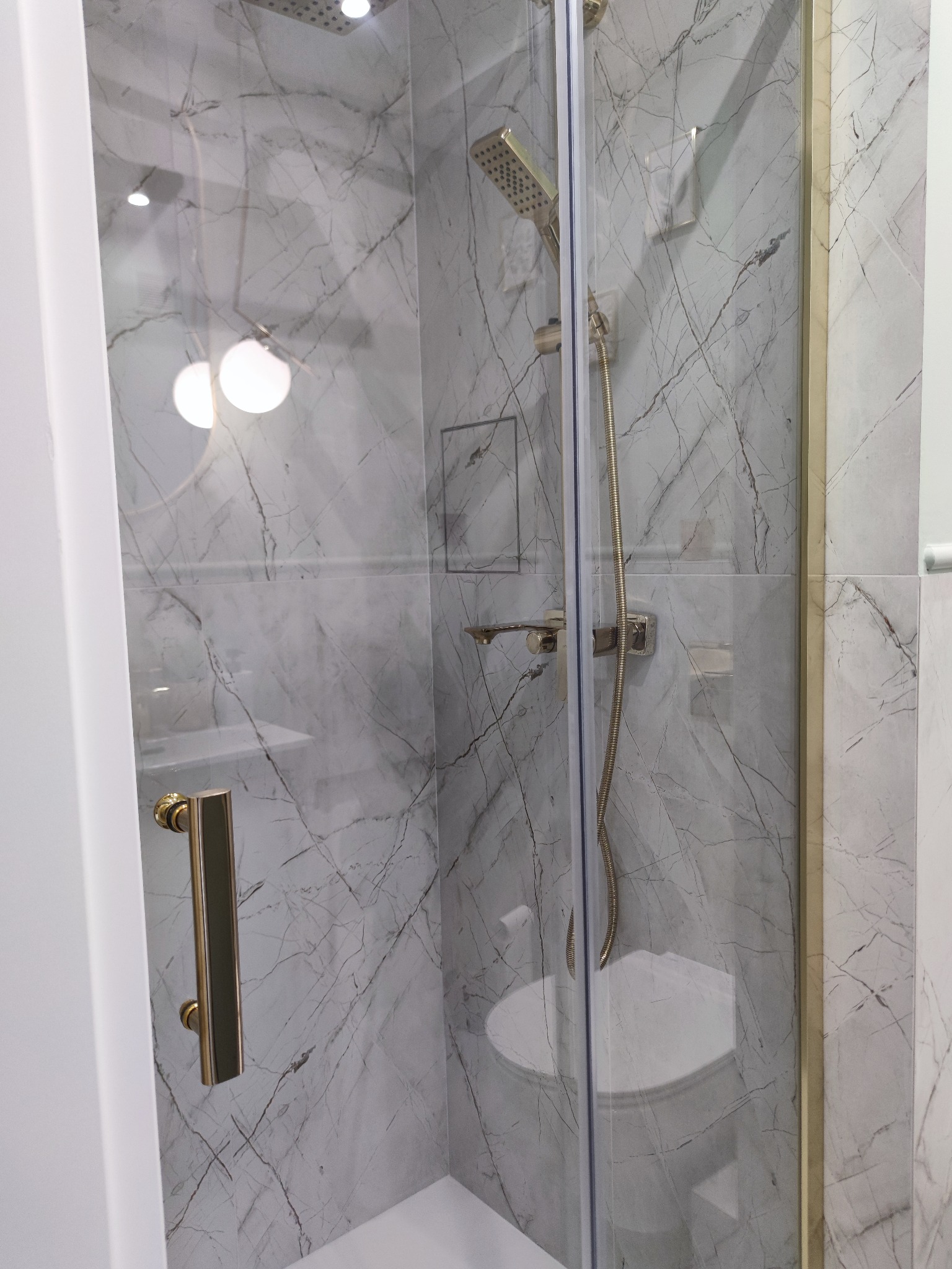 Kabina prysznicowa z marmurowymi płytkami, złotą armaturą i szklanymi drzwiami z złotym uchwytem, widok z zewnątrz.