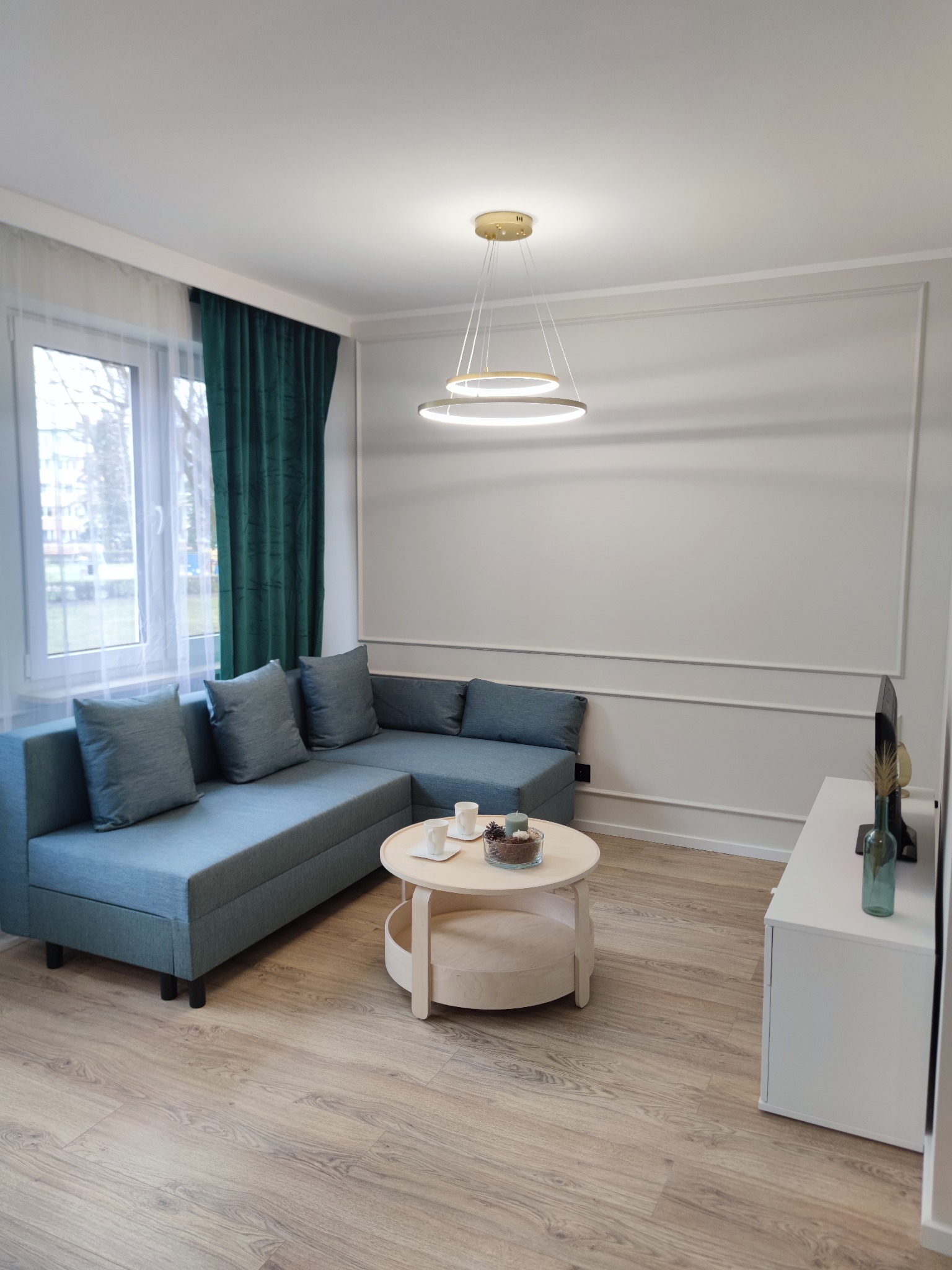 Jasne wnętrze salonu z narożną sofą w odcieniach błękitu, okrągłym stolikiem kawowym z dekoracjami, telewizorem na białej szafce RTV i nowoczesną lampą wiszącą z trzema okręgami.