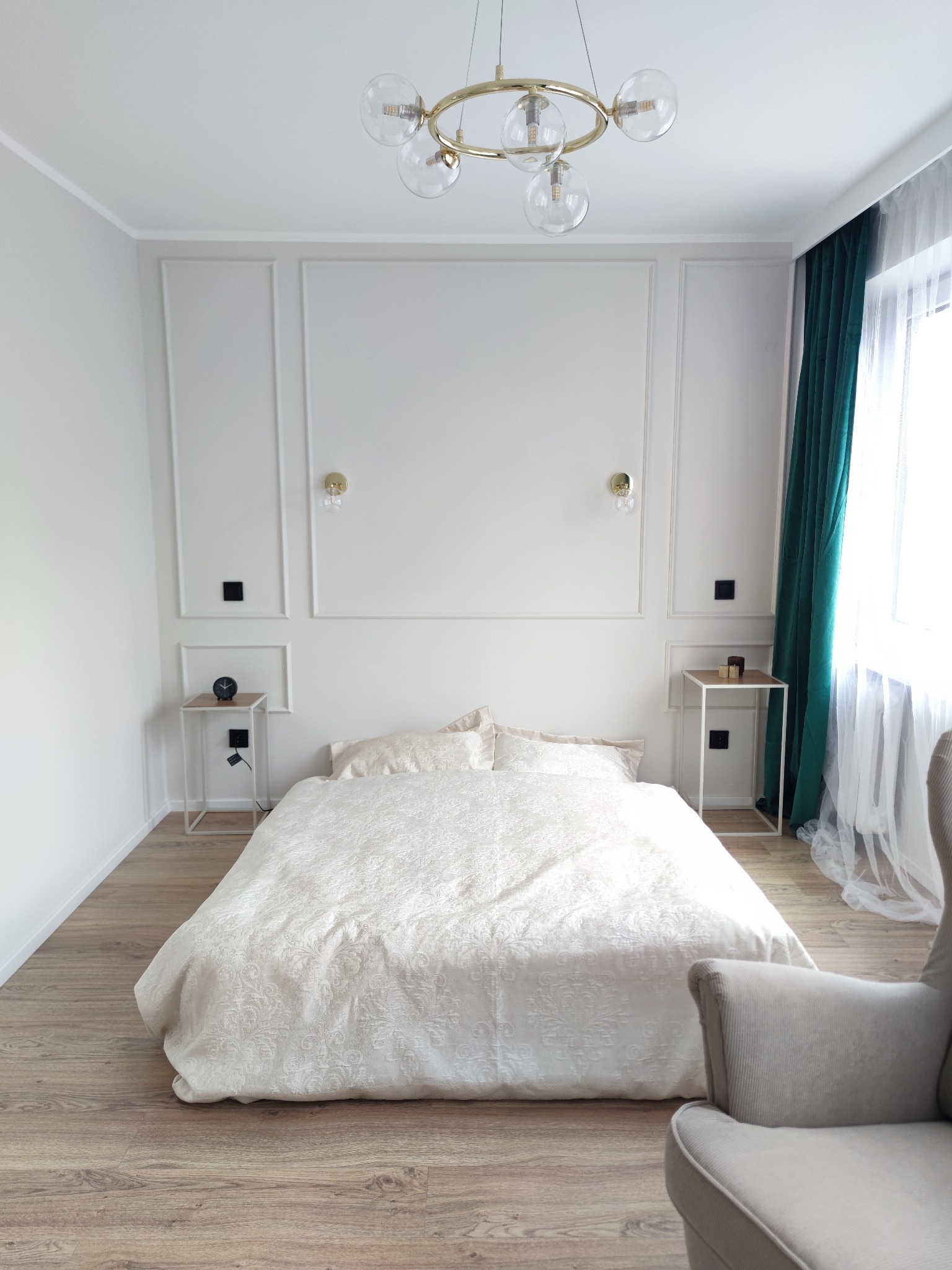 Elegancka sypialnia w jasnych barwach z dekoracyjnymi panelami ściennymi, złotymi lampkami, okrągłym żyrandolem i zasłonami w odcieniach zieleni. Na drewnianej podłodze stoi łóżko z beżową pościelą...