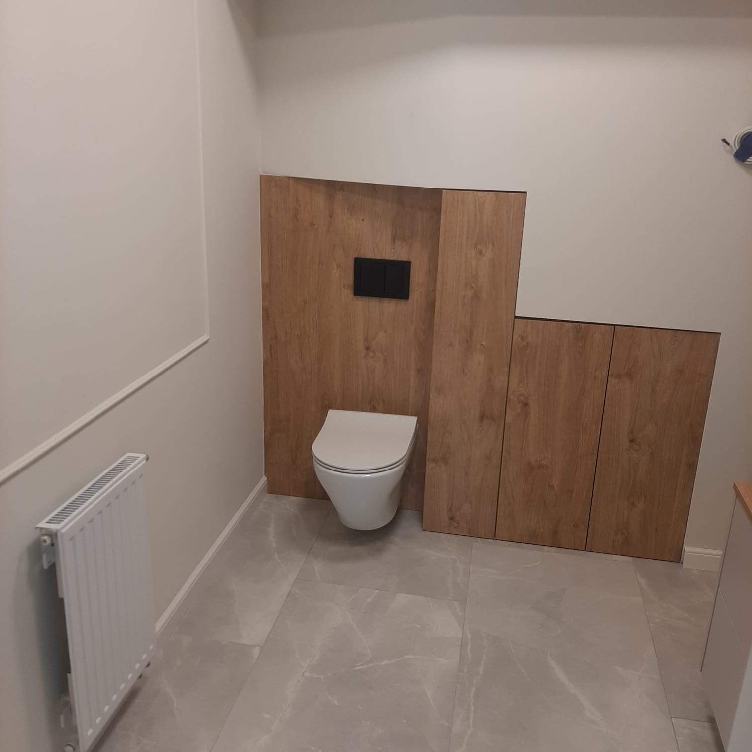 Nowoczesna łazienka z drewnianą zabudową WC, szarą marmurową podłogą i białym grzejnikiem. Minimalistyczny design i funkcjonalne rozwiązania.