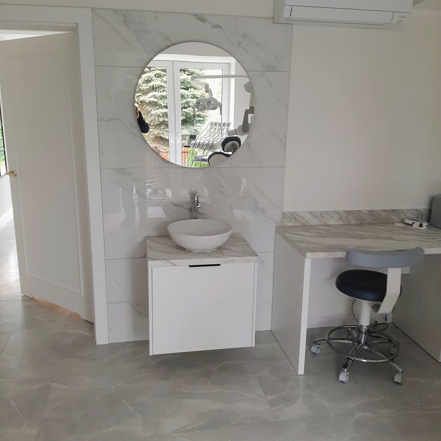 Minimalistyczna łazienka w bieli i szarości z okrągłym lustrem, marmurowym blatem i szafką pod umywalką. W tle widoczny fotel dentystyczny.