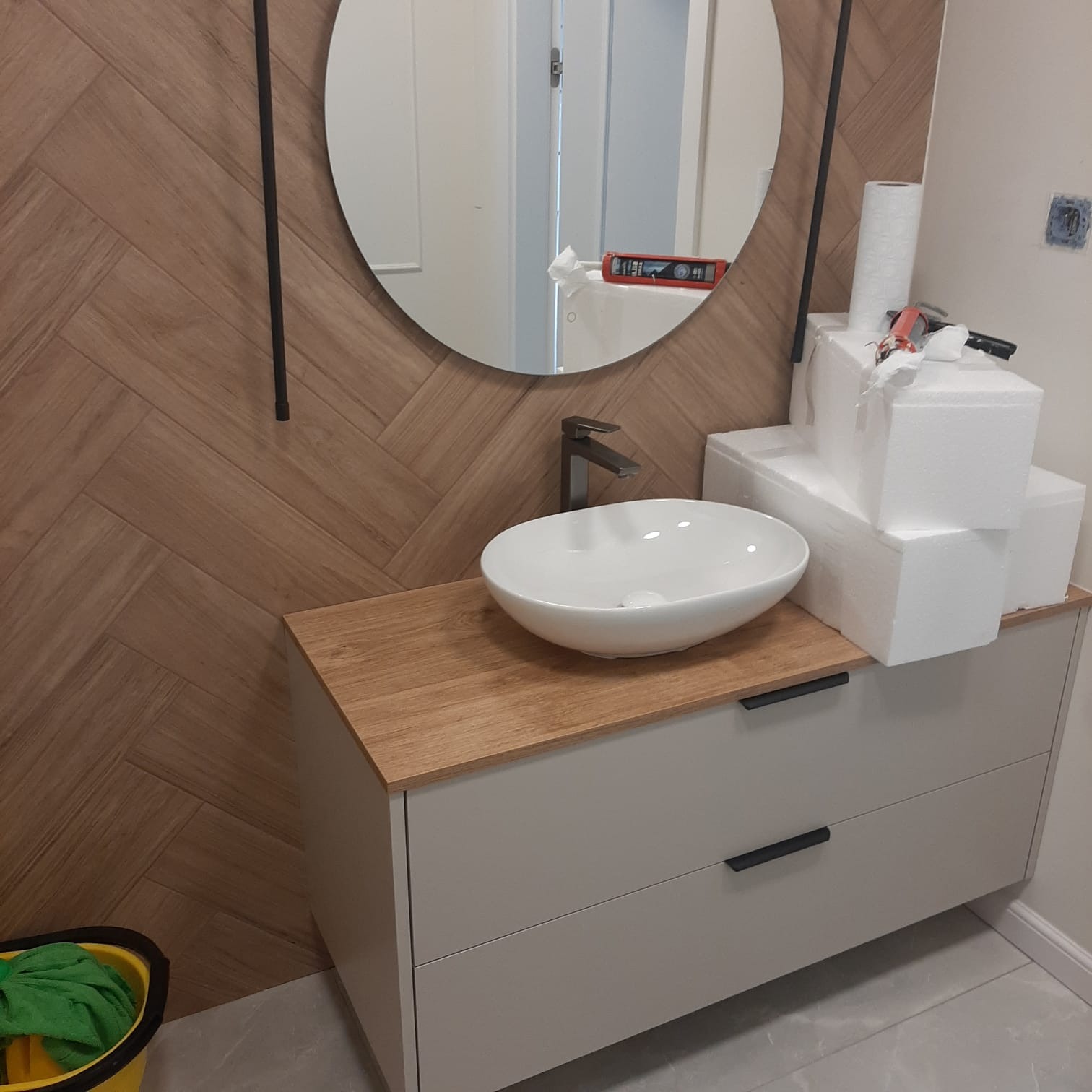 Umywalka nablatowa w nowoczesnej łazience z drewnianą ścianą w jodełkę i okrągłym lustrem. W tle akcesoria montażowe i narzędzia.