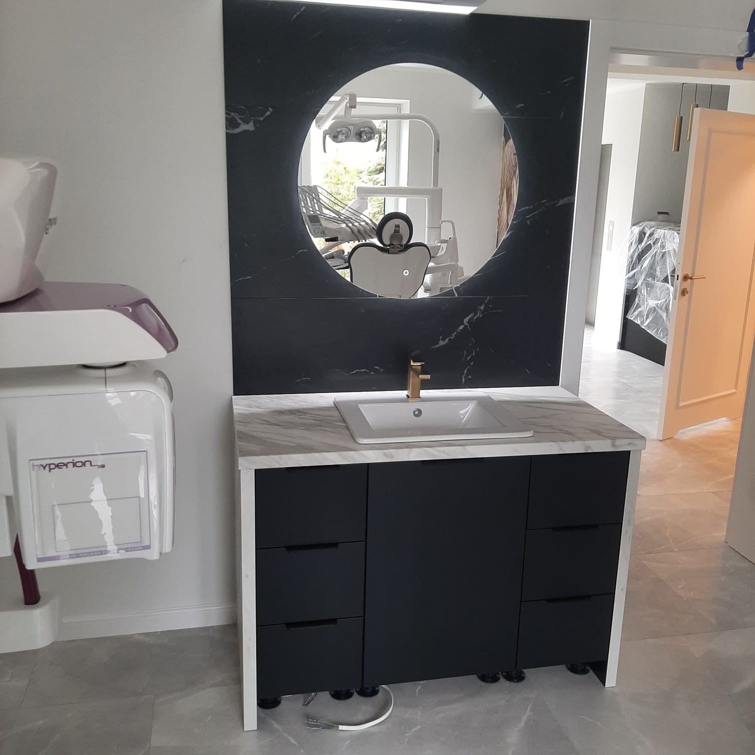 Nowoczesna łazienka z czarną szafką pod umywalką, marmurowym blatem i okrągłym lustrem, w którym odbija się fotel dentystyczny i sprzęt medyczny.