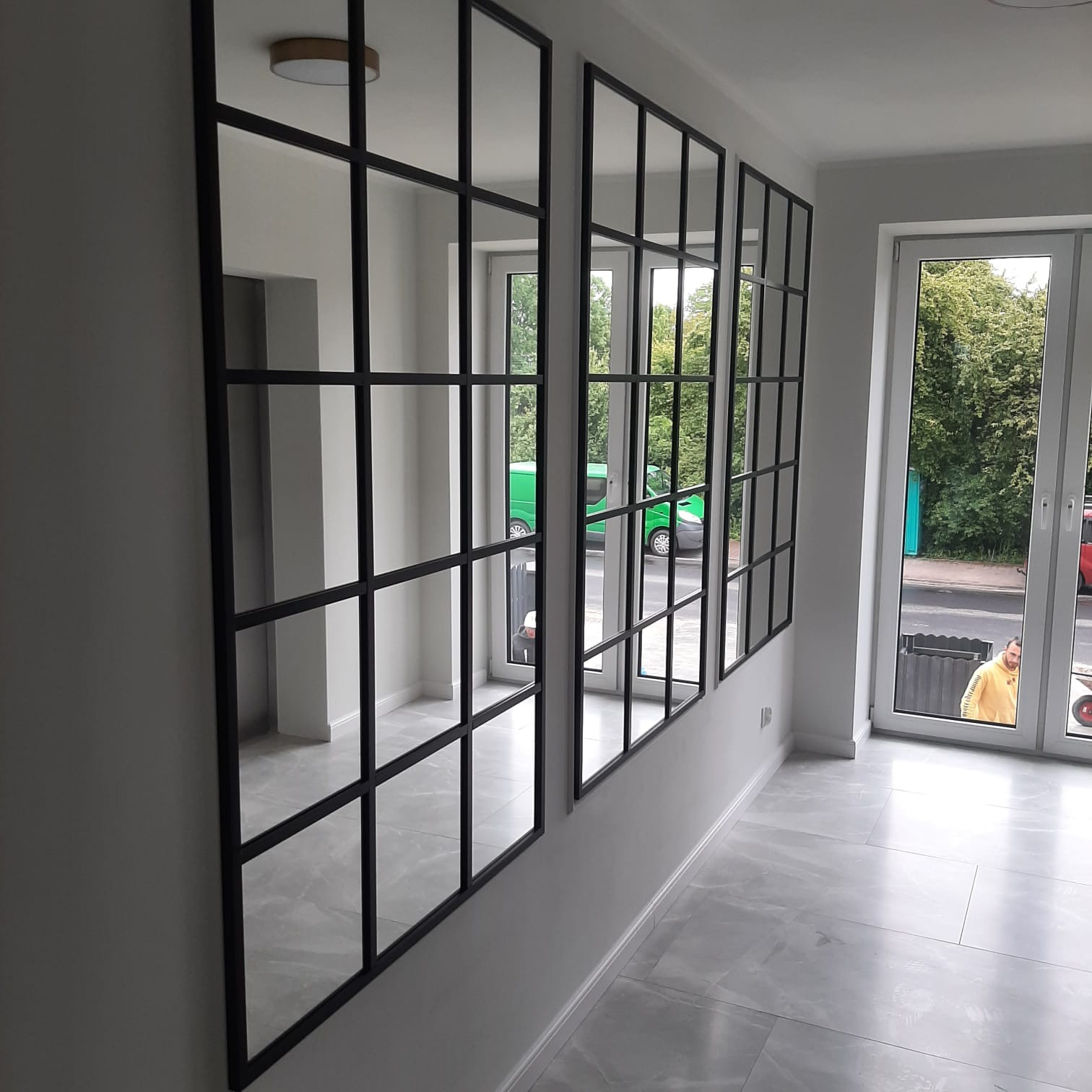 Trzy duże, czarne ramy okienne na białej ścianie, tworzące efekt lustra. Widok na szarą podłogę i okno z widokiem na ulicę. Minimalistyczny design.