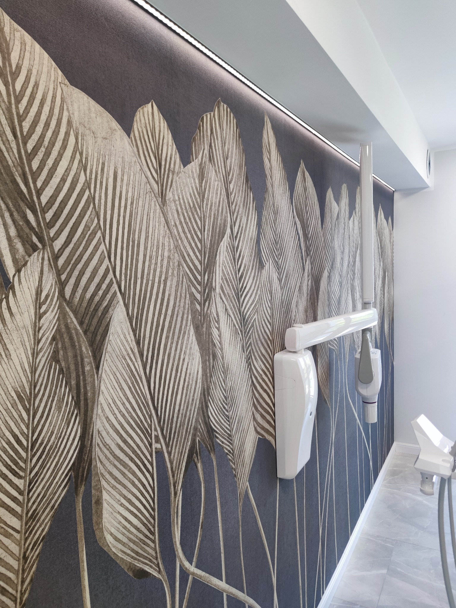 Ściana w gabinecie z tapetą w liście i nowoczesnym oświetleniem LED, uzupełniona o biały sprzęt dentystyczny. Minimalistyczny design.