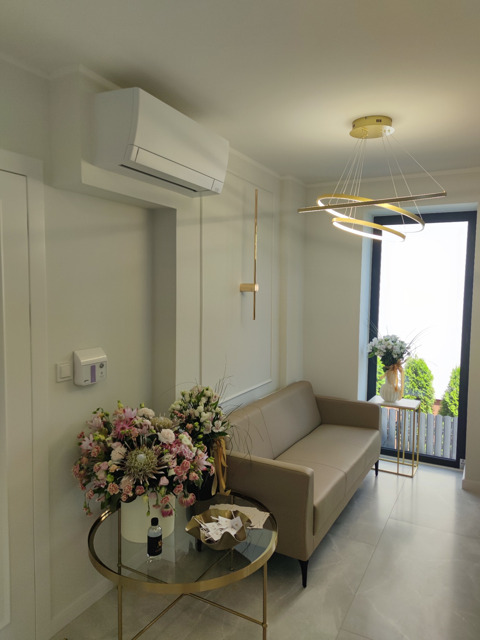 Nowoczesne wnętrze: beżowa sofa, okrągły stolik z kwiatami, designerska lampa nad oknem z widokiem na zieleń. Minimalistyczny klimatyzator na ścianie.