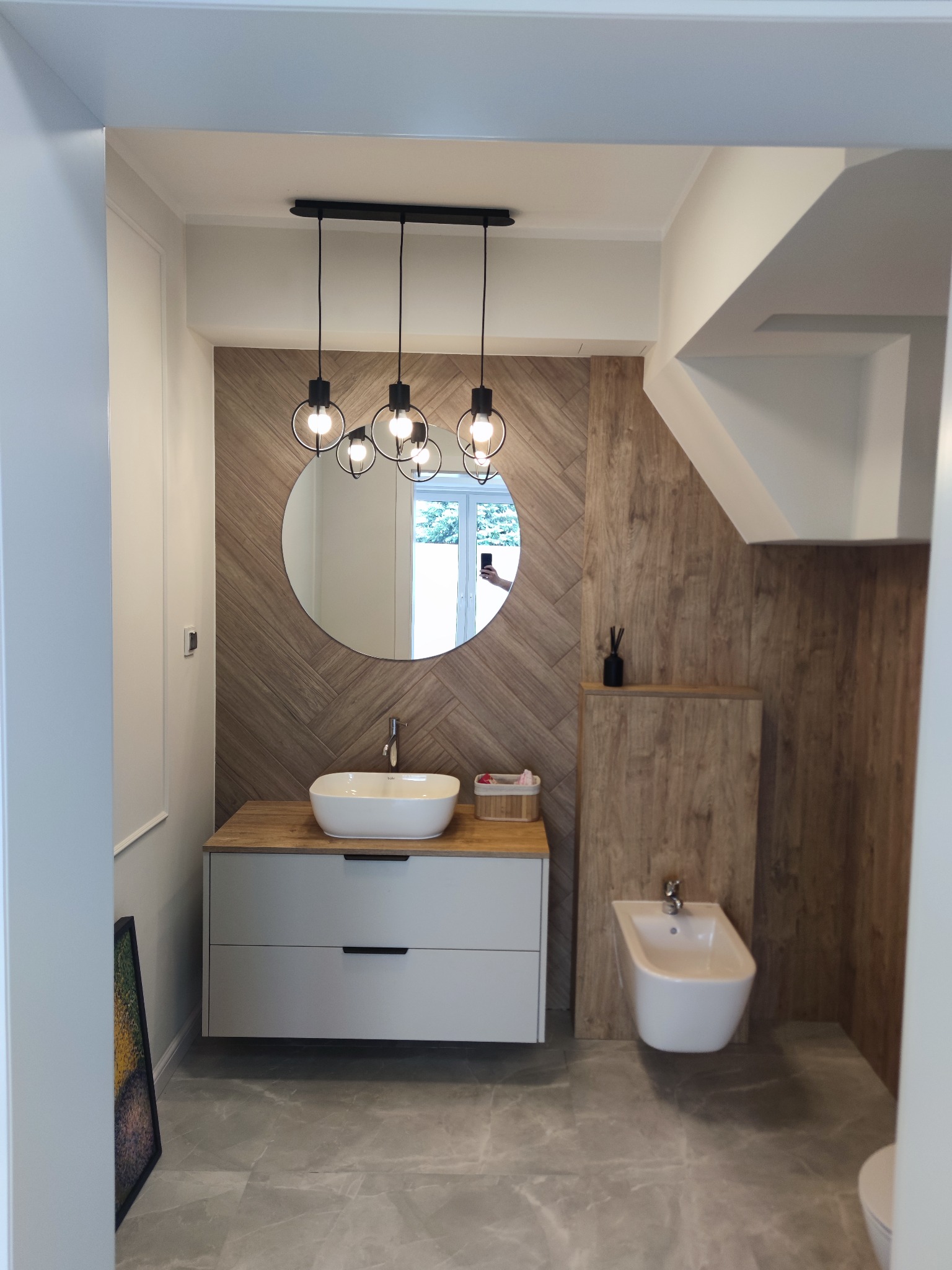 Nowoczesna łazienka z drewnianą ścianą, okrągłym lustrem i wiszącą lampą. Biała umywalka nablatowa i bidet na tle szarych płytek podłogowych.