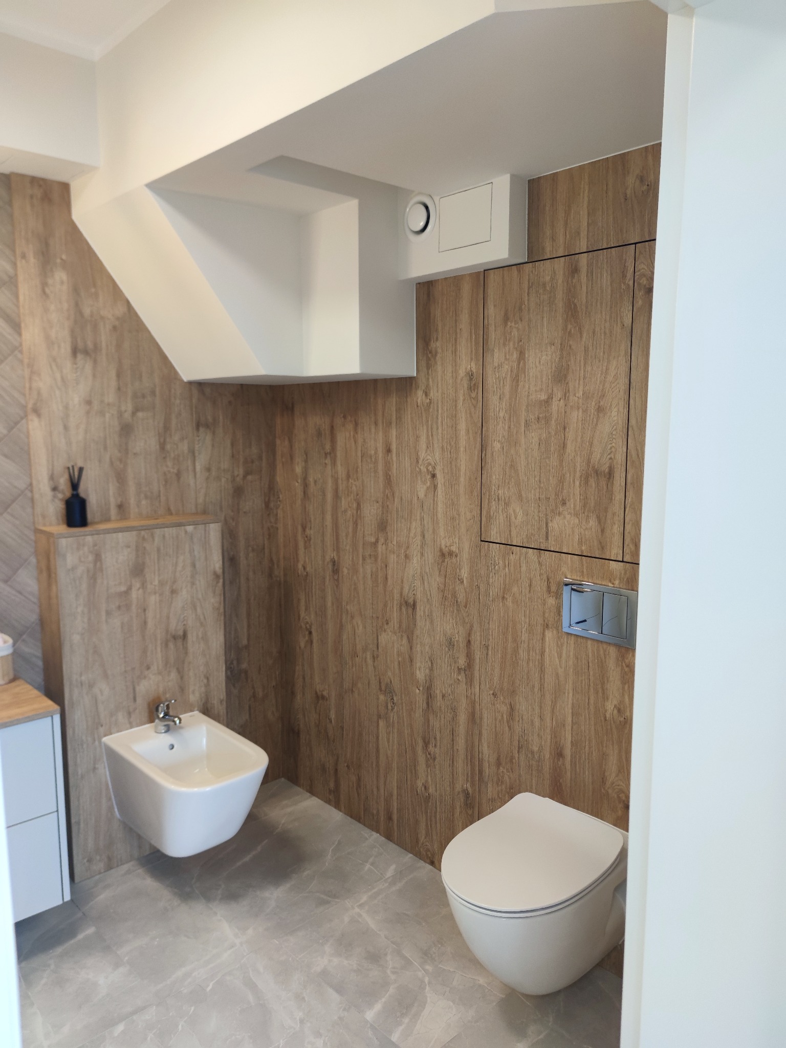 Nowoczesna łazienka z drewnopodobnymi panelami ściennymi, szarą podłogą i białymi sanitariatami: toaleta podwieszana i bidet. Ciekawy układ wentylacji pod sufitem.