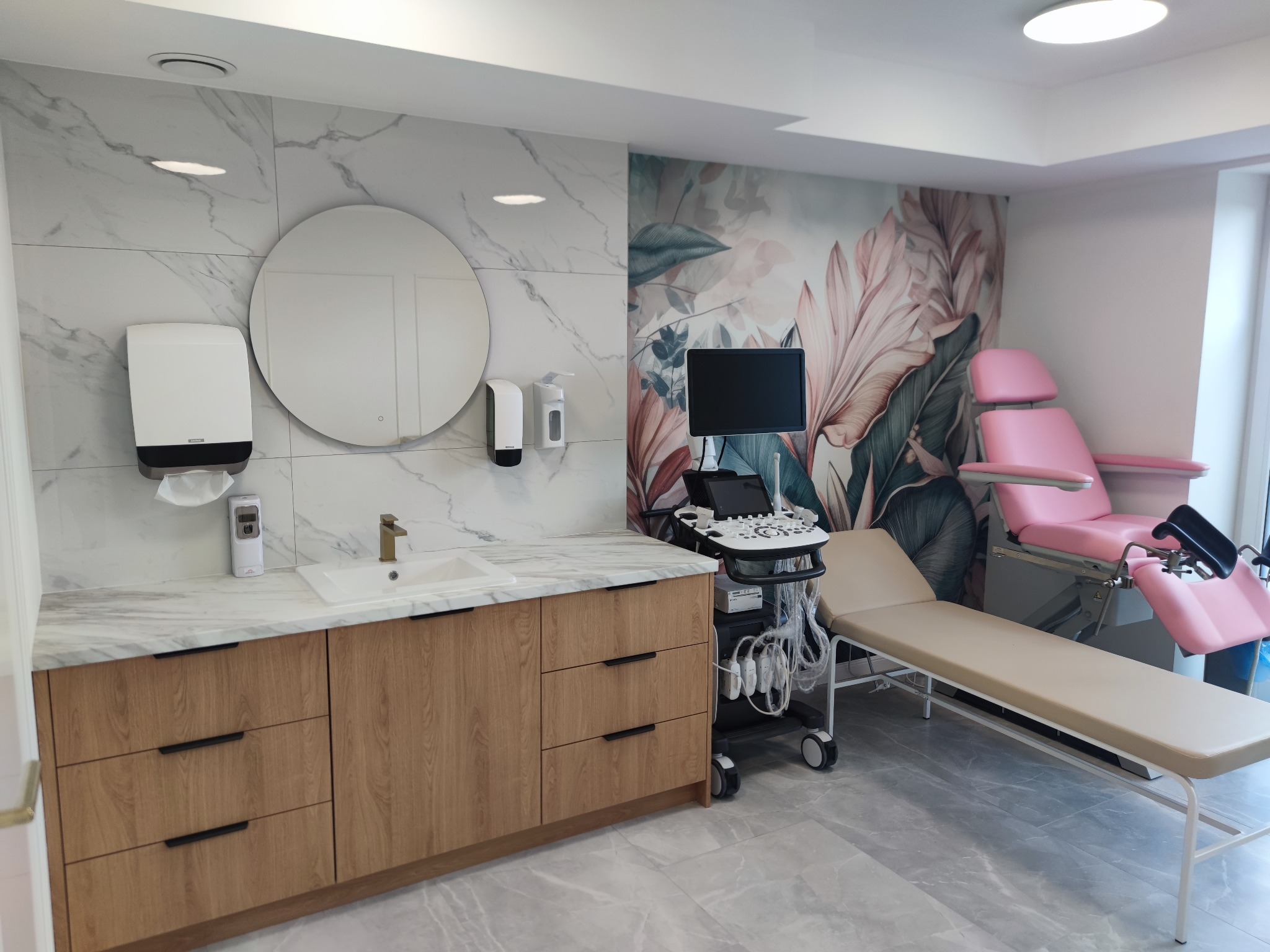 Nowoczesne wnętrze gabinetu lekarskiego z marmurową ścianą, drewnianą szafką z umywalką i sprzętem medycznym, w tym różowym fotelem ginekologicznym.