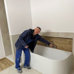 OLEKSANDR SAHIN LIDER BUD - Biała wanna w wykończonej łazience z beżowymi płytkami i drewnianymi akcentami. Instalator w ubraniu roboczym sprawdza zamontowaną armaturę.