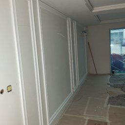 OLEKSANDR SAHIN LIDER BUD - Białe ściany z dekoracyjnymi panelami w trakcie wykańczania wnętrza. Podłoga zabezpieczona kartonem. Widoczne elementy instalacji elektrycznej.