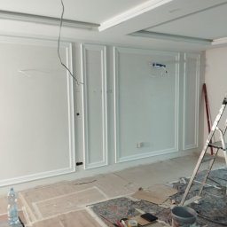 OLEKSANDR SAHIN LIDER BUD - Białe panele dekoracyjne na ścianie w trakcie montażu, widoczne narzędzia i drabina. Sufit z dekoracyjnymi wgłębieniami. Wnętrze w trakcie wykańczania.