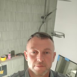 OLEKSANDR SAHIN LIDER BUD - Wykończona łazienka z szarymi, pionowymi płytkami. Widoczna kabina prysznicowa typu deszczownica i półka na kosmetyki. Na pierwszym planie twarz mężczyzny.