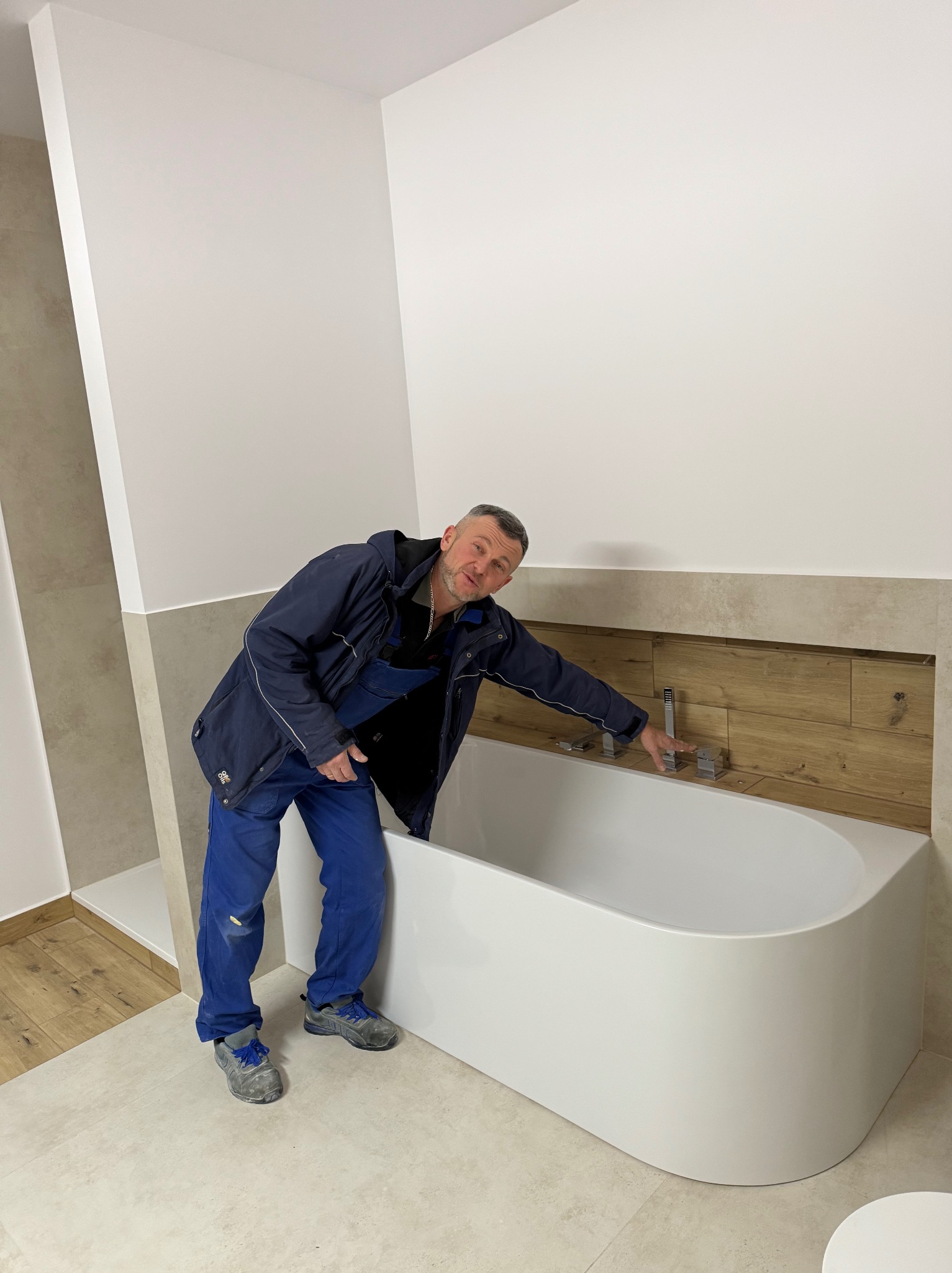 Biała wanna w wykończonej łazience z beżowymi płytkami i drewnianymi akcentami. Instalator w ubraniu roboczym sprawdza zamontowaną armaturę.