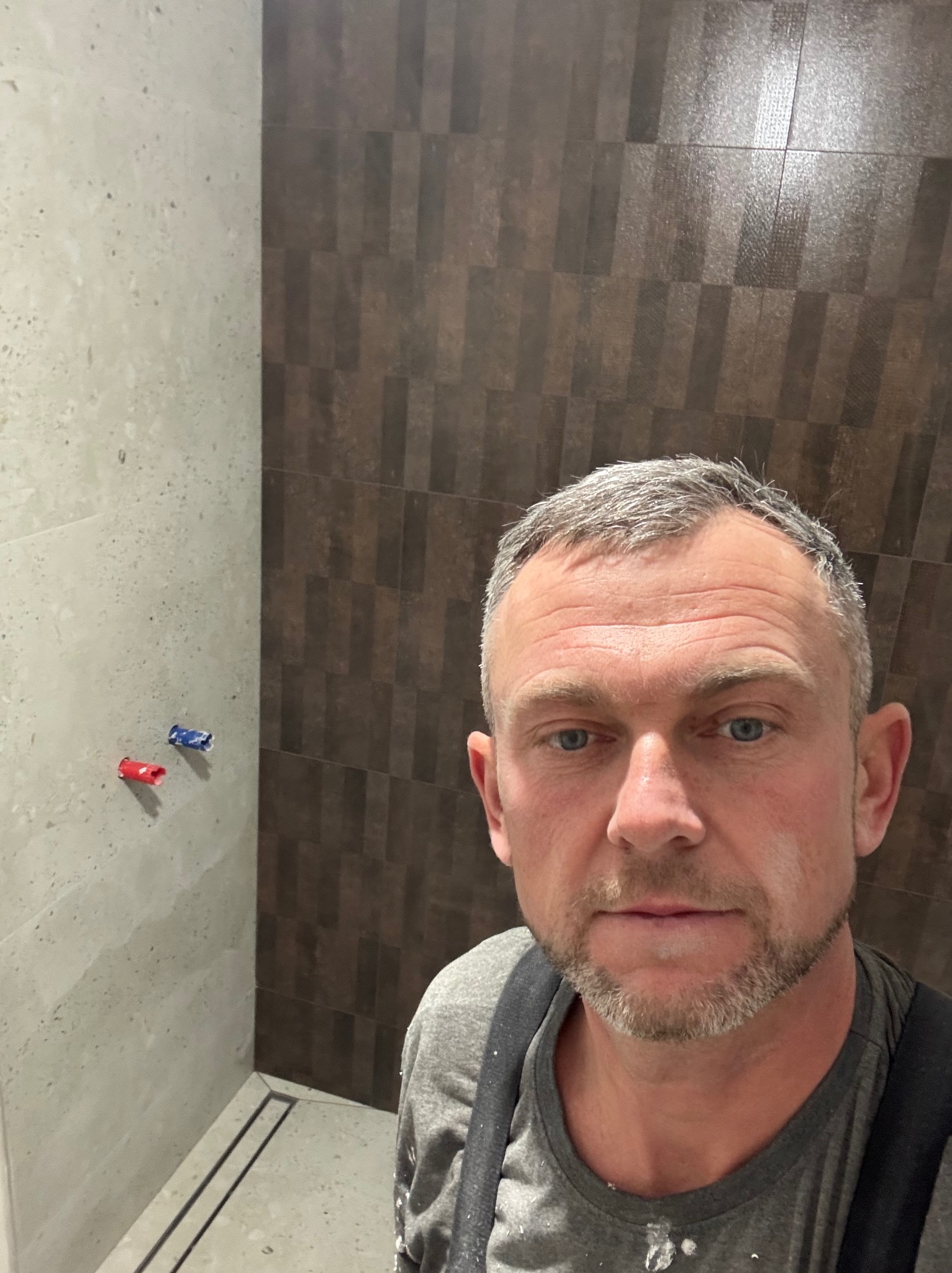 Wykończona łazienka z brązowo-beżowymi płytkami, widoczny odpływ liniowy i instalacja wodna. Mężczyzna w tle, prawdopodobnie wykonawca, robi selfie.