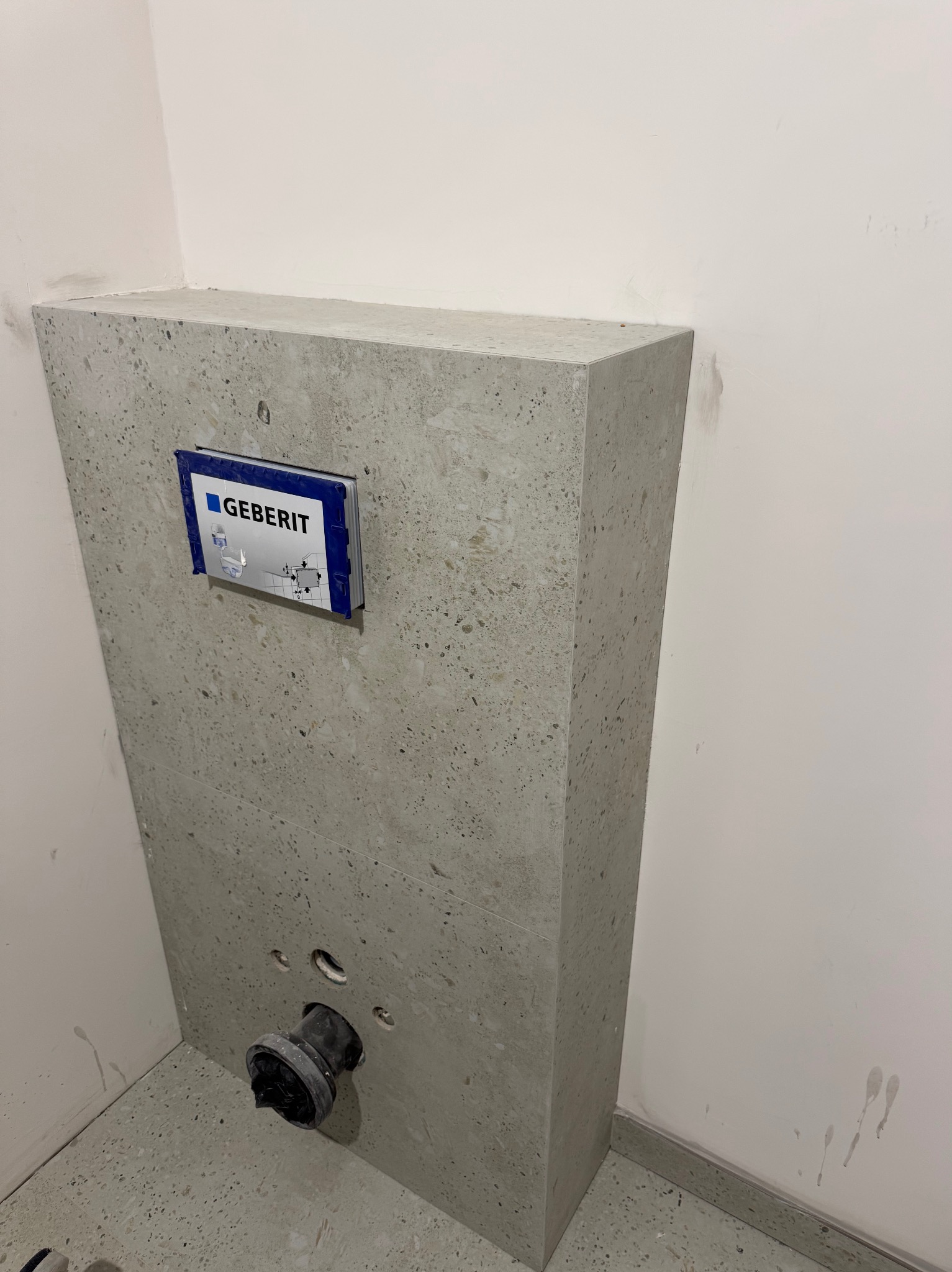 Szary, kamienny stelaż podtynkowy Geberit w łazience, z widocznymi przyłączami. Wykończenie w stylu lastryko, surowy minimalizm.