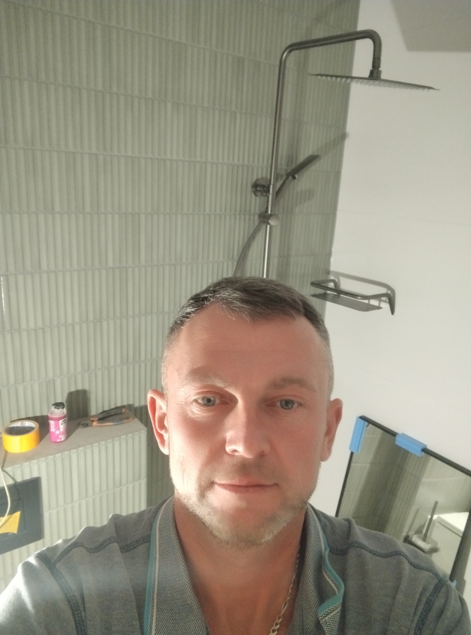 Wykończona łazienka z szarymi, pionowymi płytkami. Widoczna kabina prysznicowa typu deszczownica i półka na kosmetyki. Na pierwszym planie twarz mężczyzny.