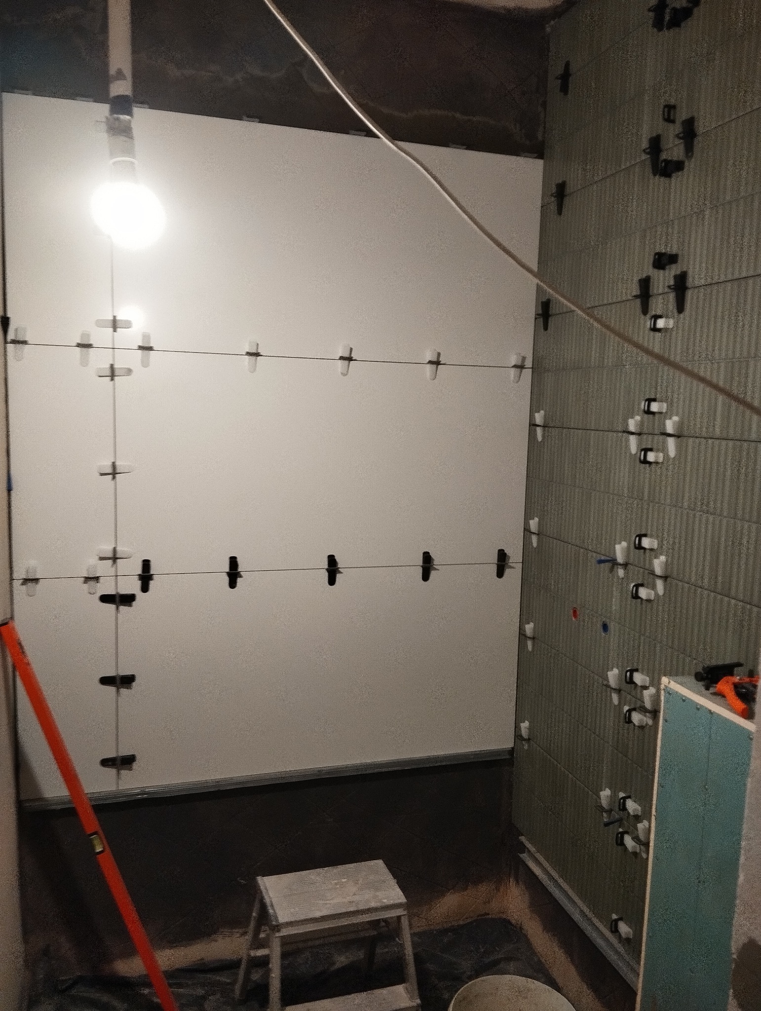 Ściana łazienki w trakcie układania płytek, widoczne krzyżyki dystansowe i poziomica. Częściowo wykończona ściana z szarymi płytkami, druga w trakcie prac z białymi płytkami.