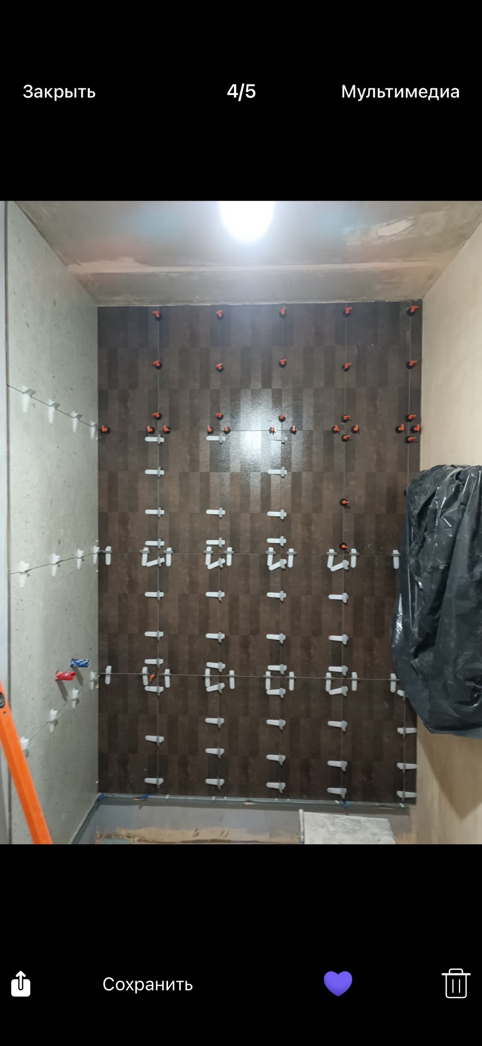 Układanie brązowych płytek w szachownicę na ścianie łazienki z użyciem krzyżyków dystansowych i poziomicy. Widoczny fragment podłogi i zabezpieczona folią wanna.