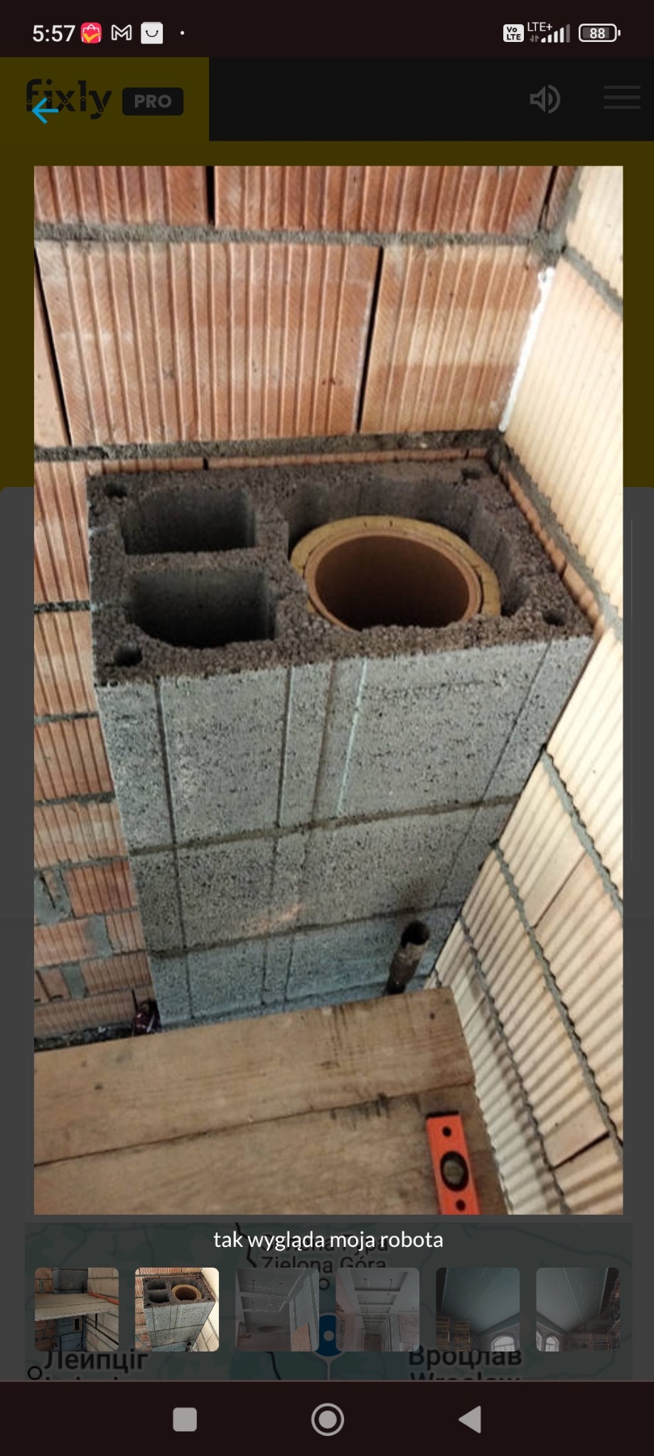 Wznoszenie komina z szarego bloczka betonowego, widoczne elementy ceramiczne wewnątrz. Poziomica na drewnianej desce. Ceglana ściana w tle.