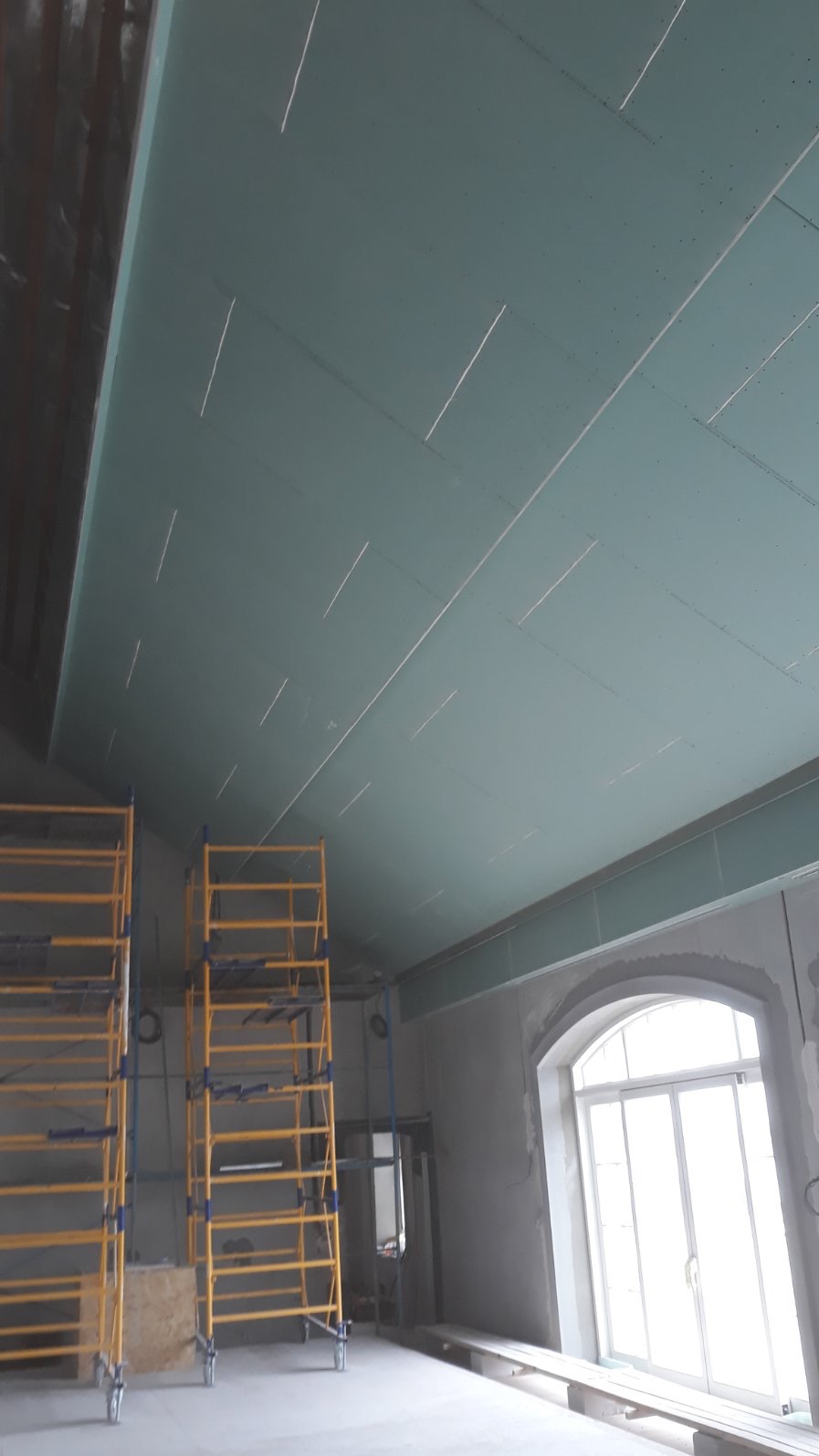 Montaż płyt karton gips na suficie podwieszanym. Widoczne rusztowania i okno. Etap prac wykończeniowych w pomieszczeniu.