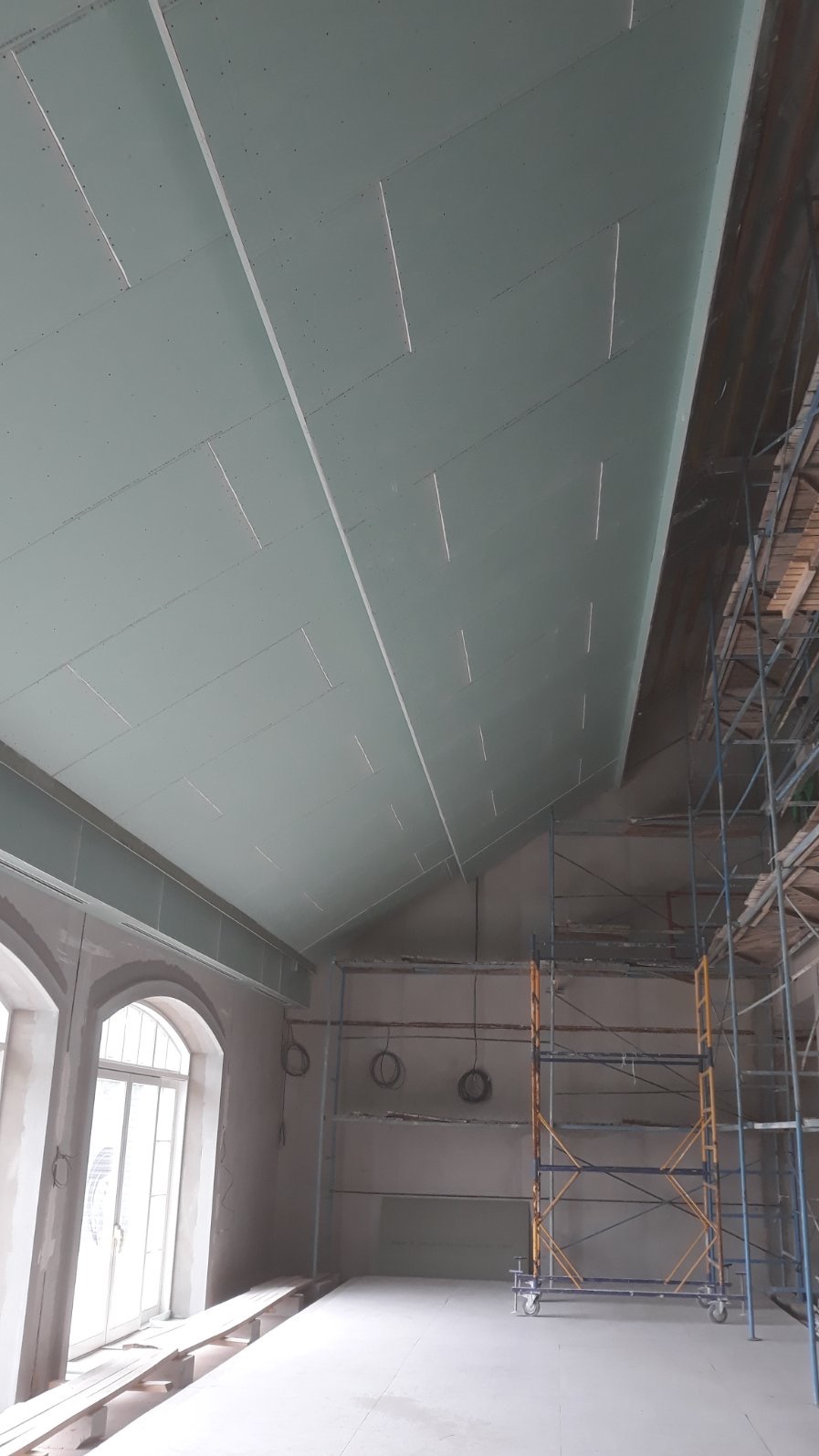 Sufit z płyt kartonowo-gipsowych w trakcie montażu, widoczne łączenia i rusztowanie. Duże okna łukowe w tle. Prace wykończeniowe w przestronnym wnętrzu.