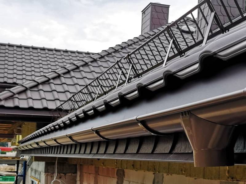 Nowy, brązowy dach z zainstalowanymi rynnami i zabezpieczeniem przeciwśniegowym, widok od dołu na tle częściowo ocieplonej ściany budynku.