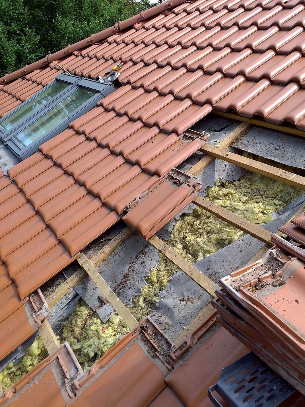 Demontaż czerwonej dachówki przy oknie dachowym, widoczna izolacja i konstrukcja drewniana dachu.