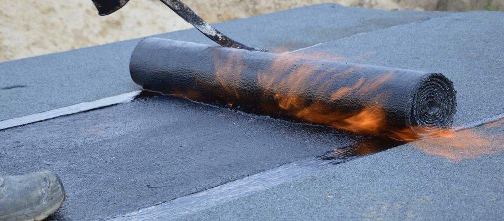 Układanie papy termozgrzewalnej na dachu za pomocą palnika, widoczny płomień i rolka materiału.