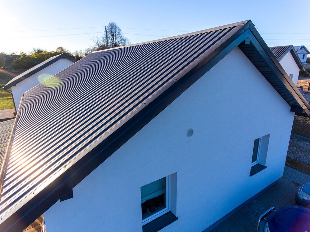 Nowoczesny dom z ciemnoszarą blachodachówką trapezową i białymi ścianami, widok z góry, w tle inne budynki.