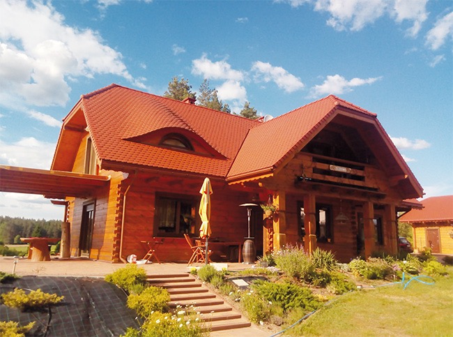 Nowy, czerwony dach z dachówki bitumicznej na drewnianym domu z bali, widok z przodu, niebieskie niebo z chmurami w tle.