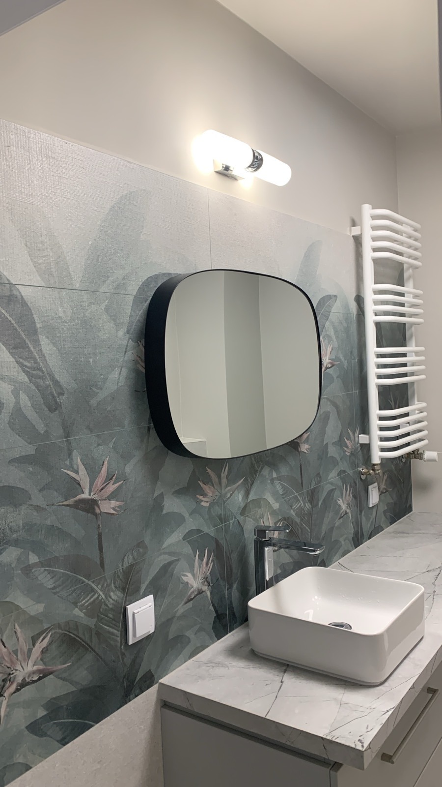 Nowoczesna łazienka z płytkami w motywy tropikalne, owalnym lustrem w czarnej ramie, białą umywalką nablatową i grzejnikiem drabinkowym.