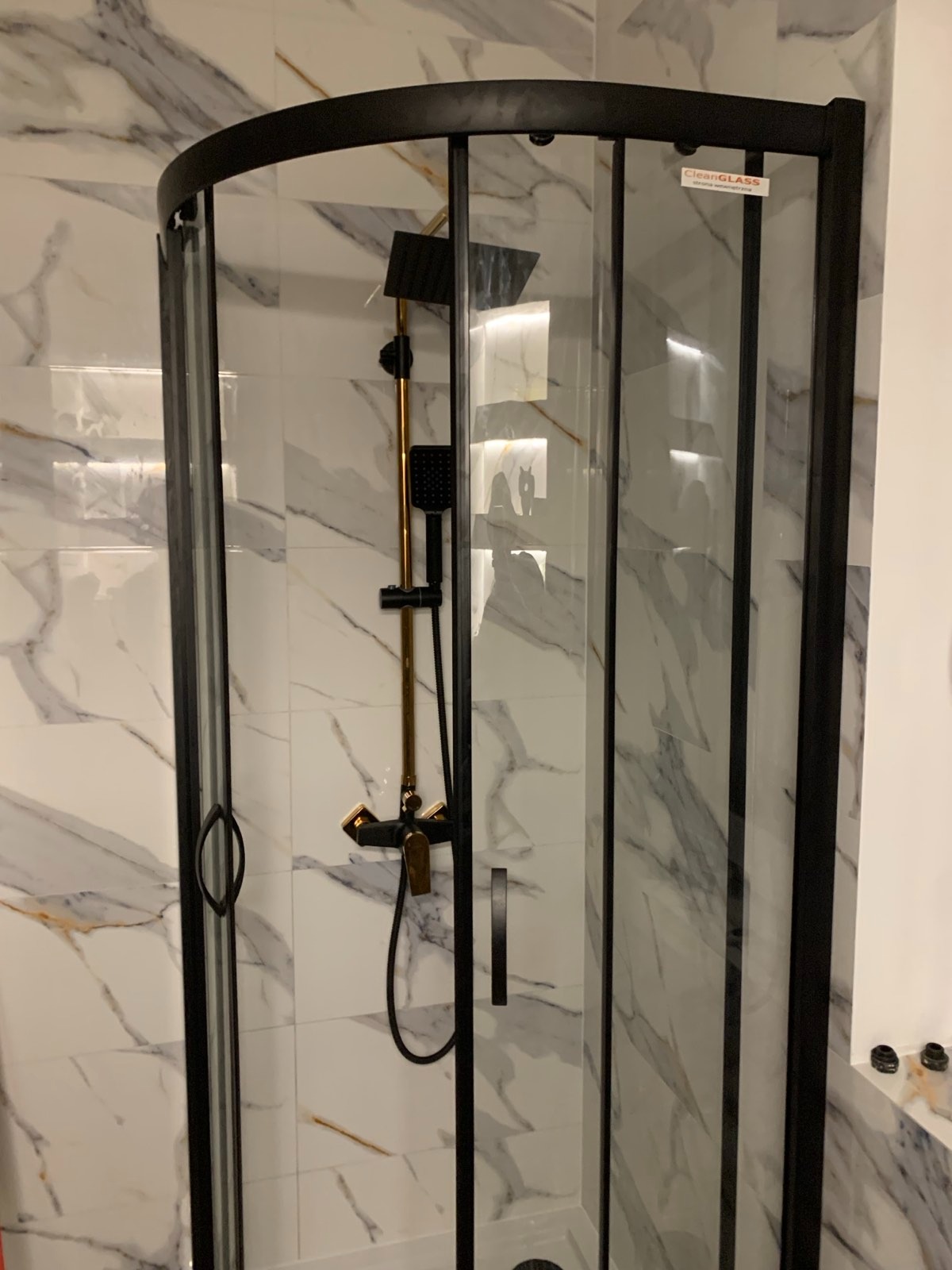Półokrągła kabina prysznicowa z czarną ramą i złotą armaturą na tle białych płytek imitujących marmur, widoczna naklejka CleanGLASS na szybie.