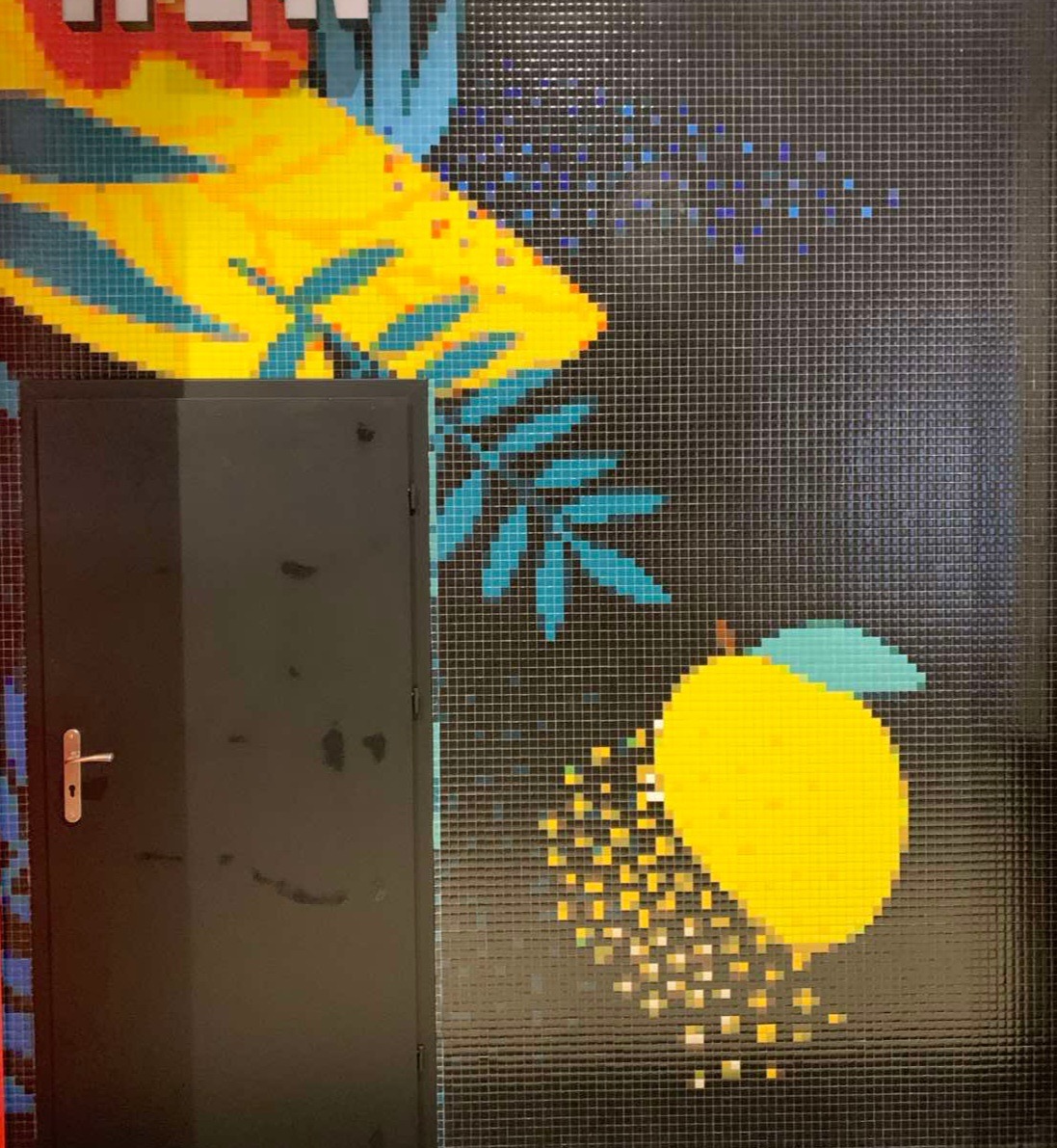 Mozaika na ścianie z motywem owoców i roślin, sąsiadująca z czarnymi drzwiami z klamką.