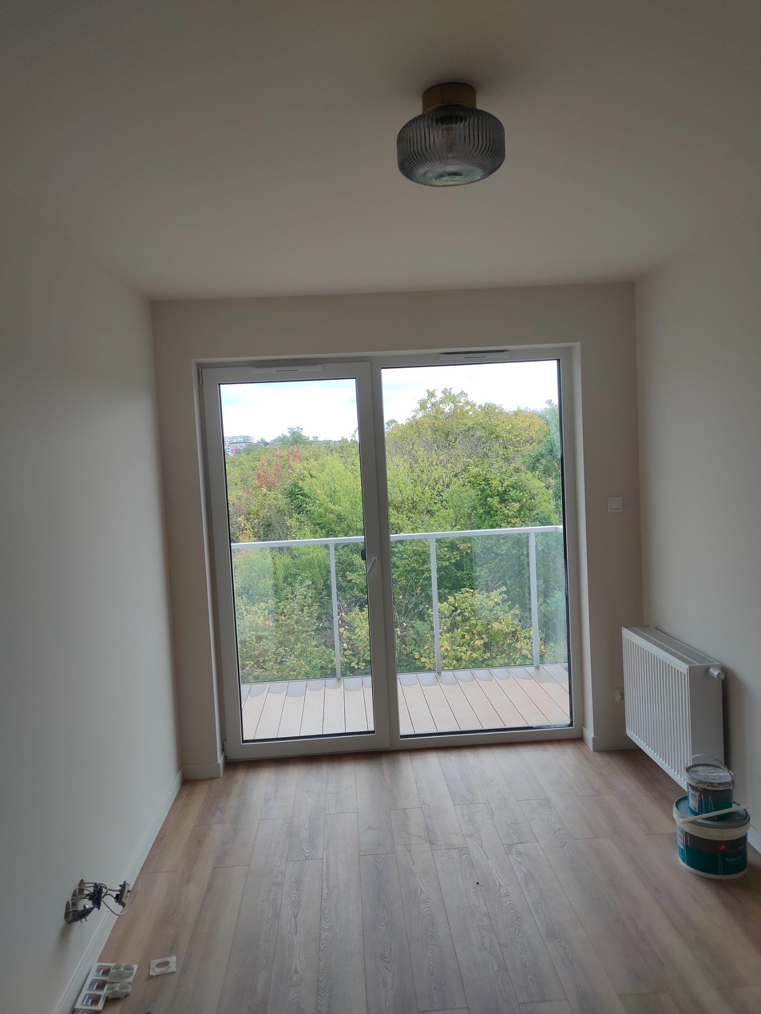 Pomalowane na biało wnętrze pokoju z parkietem, grzejnikiem i drzwiami balkonowymi z widokiem na zieleń, widoczne puszki z farbą