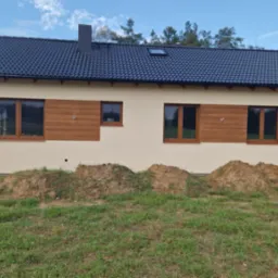 Nowo wybudowany dom jednorodzinny z beżową elewacją, ciemną dachówką i brązowymi oknami, widoczny z poziomu trawnika z usypanymi kopcami ziemi.